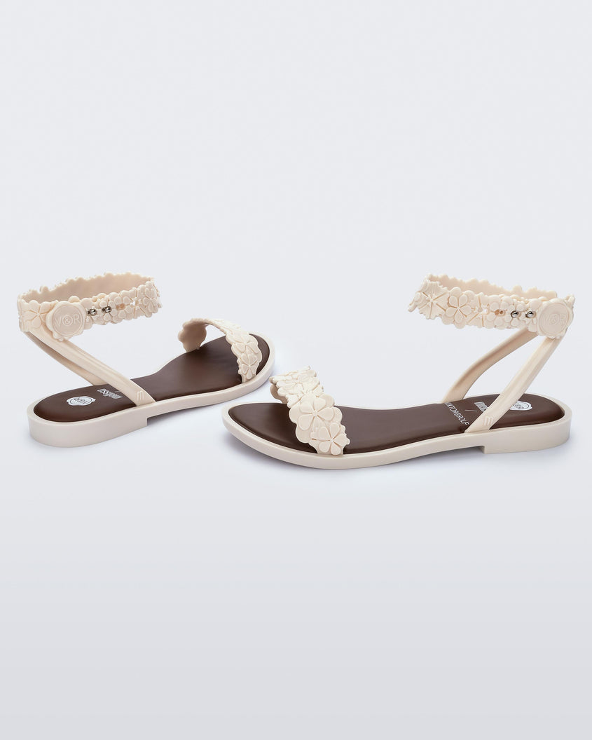 An angled back and side view of a pair of beige/brown Melissa Wave Blossom Sandals with a floral detail front and ankle strap.