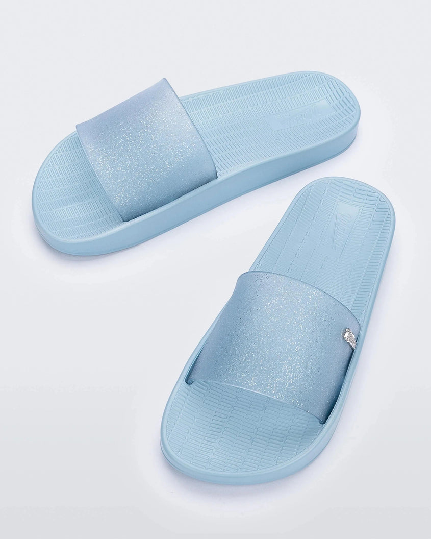 Top view of a pair of blue/clear glitter Melissa Sun Sunset slides with a blue sole and clear blue glitter front strap.