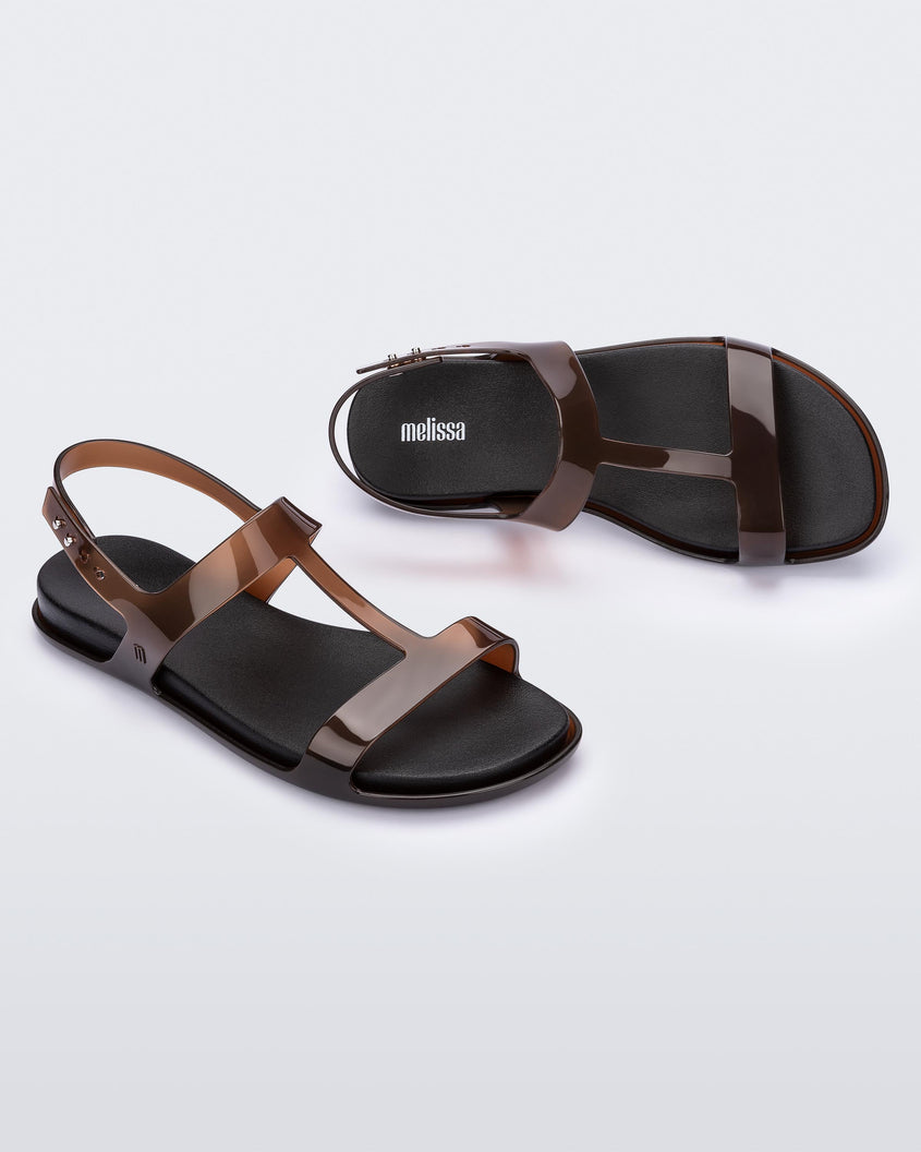 An angled top and side view of a pair of clear brown / black Melissa Adore sandals with two translucent brown straps, joined together by a third strap and a black insole.