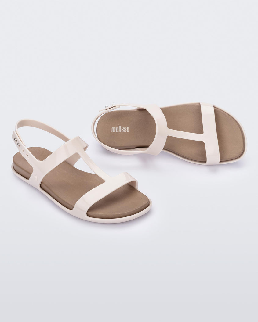 An angled top and side view of a pair of beige / brown Melissa Adore sandals with two beige straps, joined together by a third strap and a brown insole.
