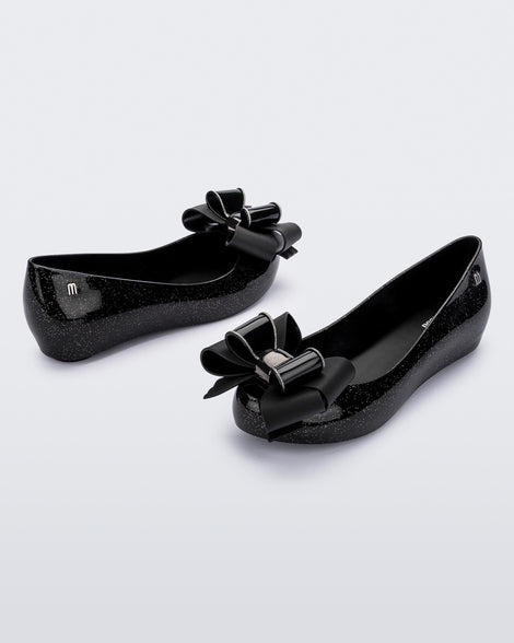 Angled view of a pair of Melissa Ultragirl ballet flat in black glitter. There is a silver M  located on the back heel. The front toe of the flat has a 3D Bow applique