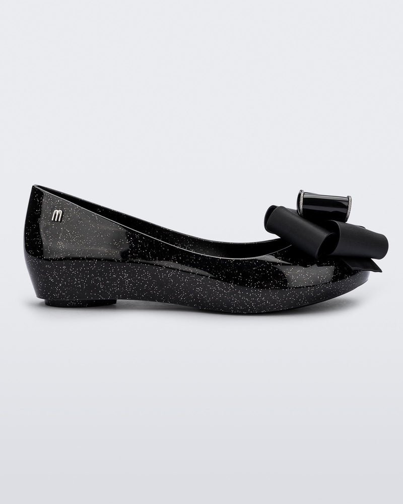 Side view of the Melissa Ultragirl ballet flat in black glitter. There is a silver M  located on the back heel. The front toe of the flat has a 3D Bow applique