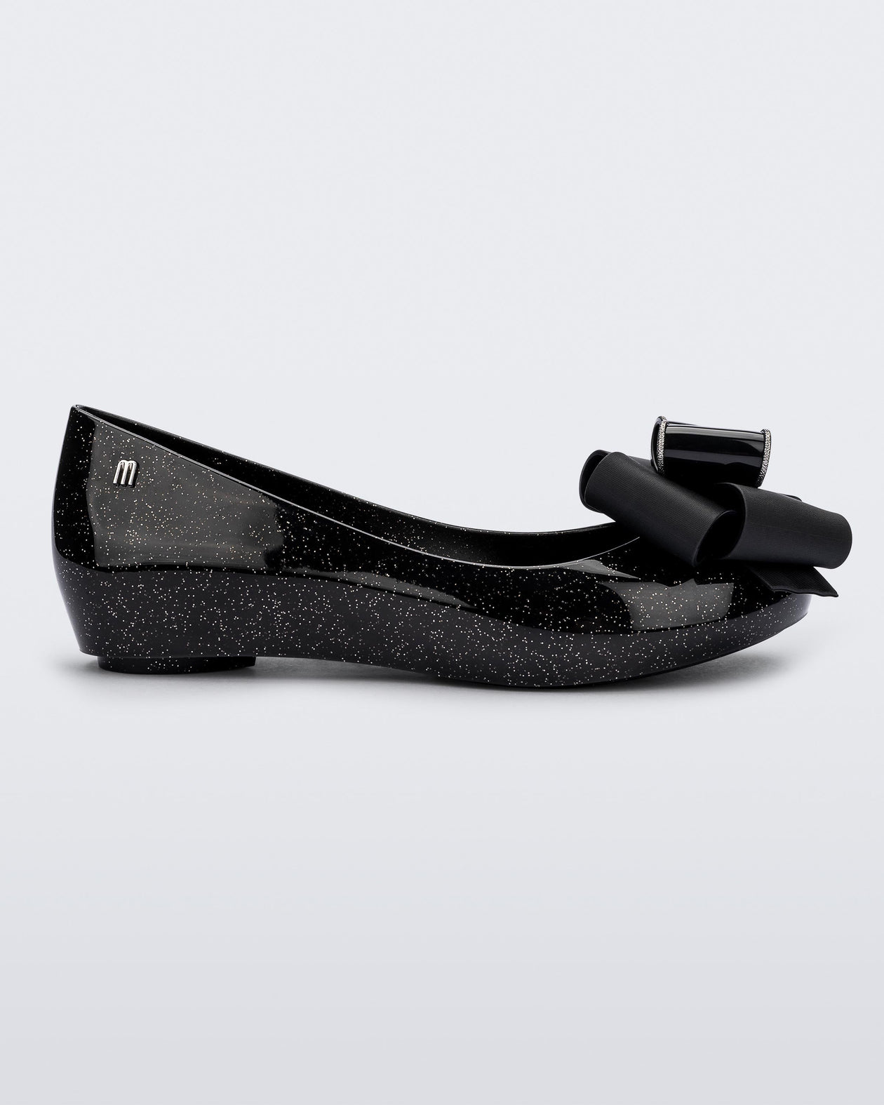Side view of the Melissa Ultragirl ballet flat in black glitter. There is a silver M  located on the back heel. The front toe of the flat has a 3D Bow applique