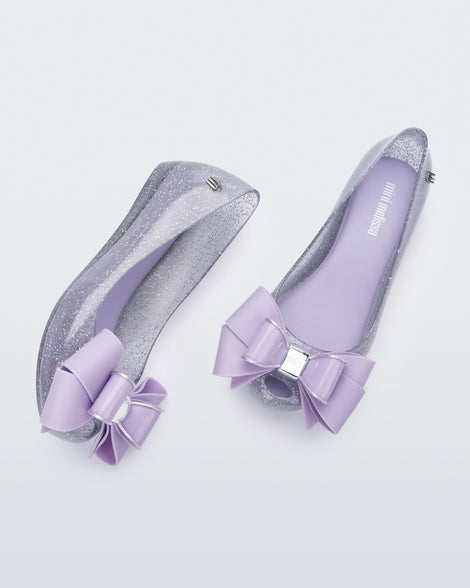 A top and side view of a pair of Clear Glitter/Lilac Mini Melissa Ultragirl Sweet flats with a clear glitter base and a lilac bow detail on the toe.