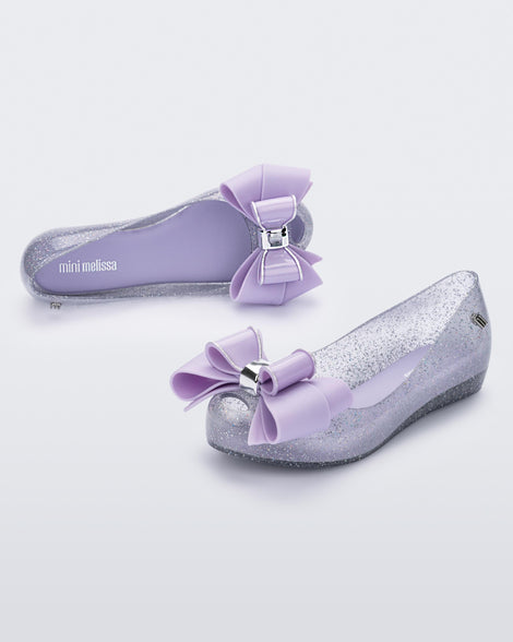 An angled front and side view of a pair of Clear Glitter/Lilac Mini Melissa Ultragirl Sweet flats with a clear glitter base and a lilac bow detail on the toe.