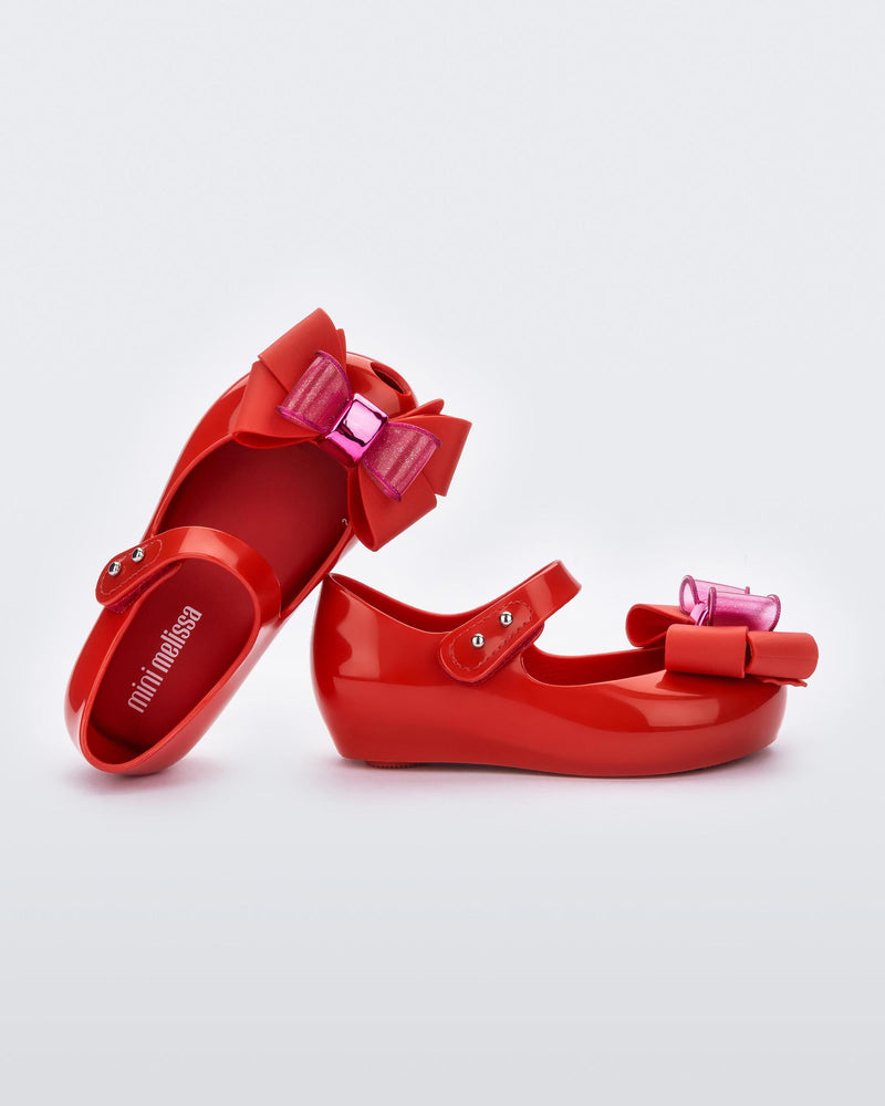 An angled front and side view of a pair of red Mini Melissa Ultragirl Sweet flats with a top strap and a pink and red bow detail on the toe, leaning on one another.