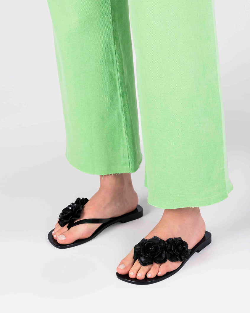 A model's legs wearing green pants and a pair of black Melissa Harmomic Squared Garden flip flops with flowers on the straps.