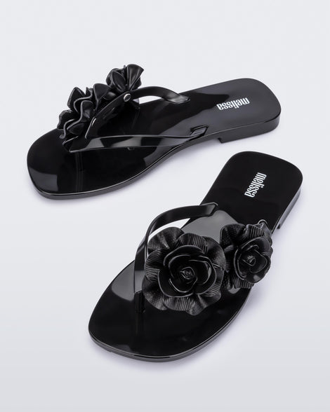An angled front view of a pair of black Melissa Harmomic Squared Garden flip flops with flowers on the straps.