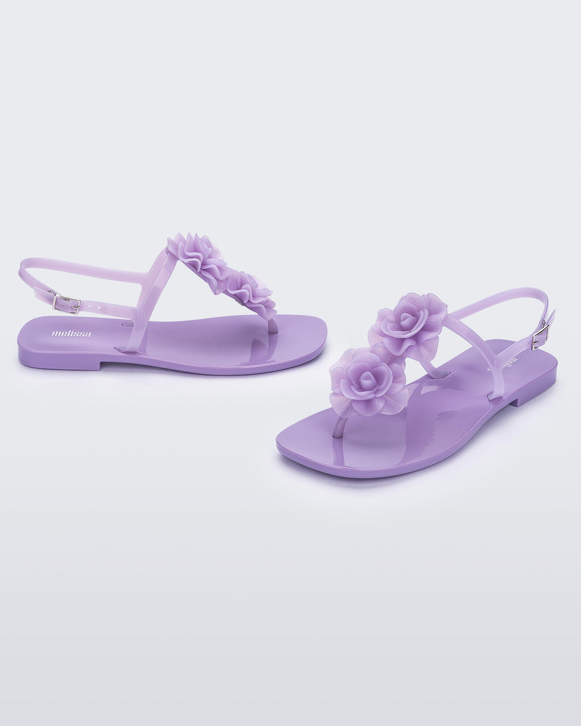 An angled front and side view of a pair of lilac Melissa Harmonic Squared Garden sandals with flower decorations on the front straps.