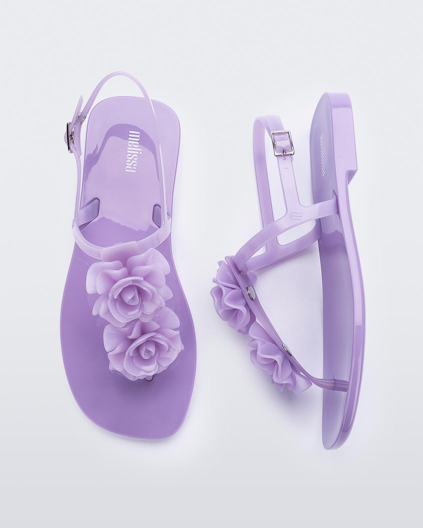 A top and side view of a pair of lilac Melissa Harmonic Squared Garden sandals with flower decorations on the front straps.