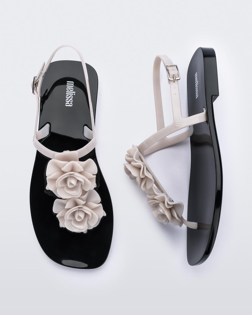 A top and side view of a pair of beige/black Melissa Harmonic Squared Garden sandals with a beige flower decoration on the front strap and a black sole.
