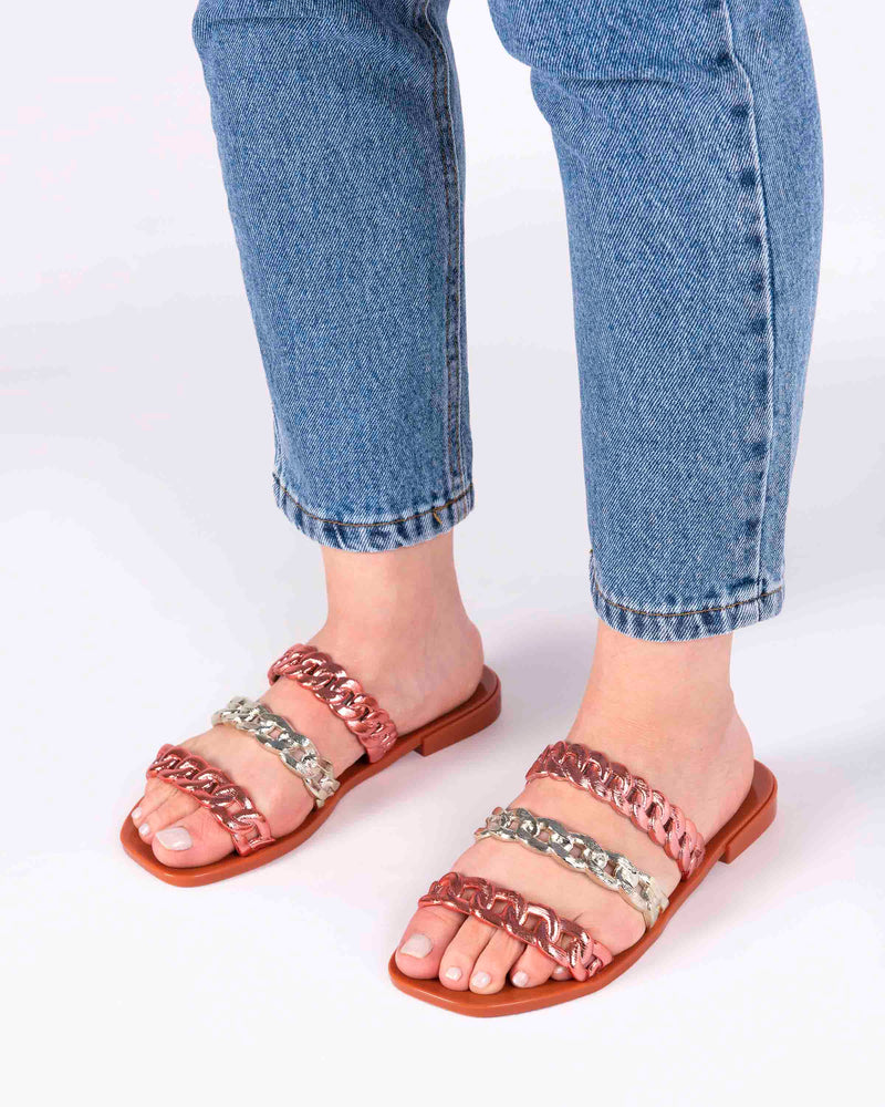 A model's legs wearing a pair of orange Melissa Feel slides with three orange and gold chain detail straps.