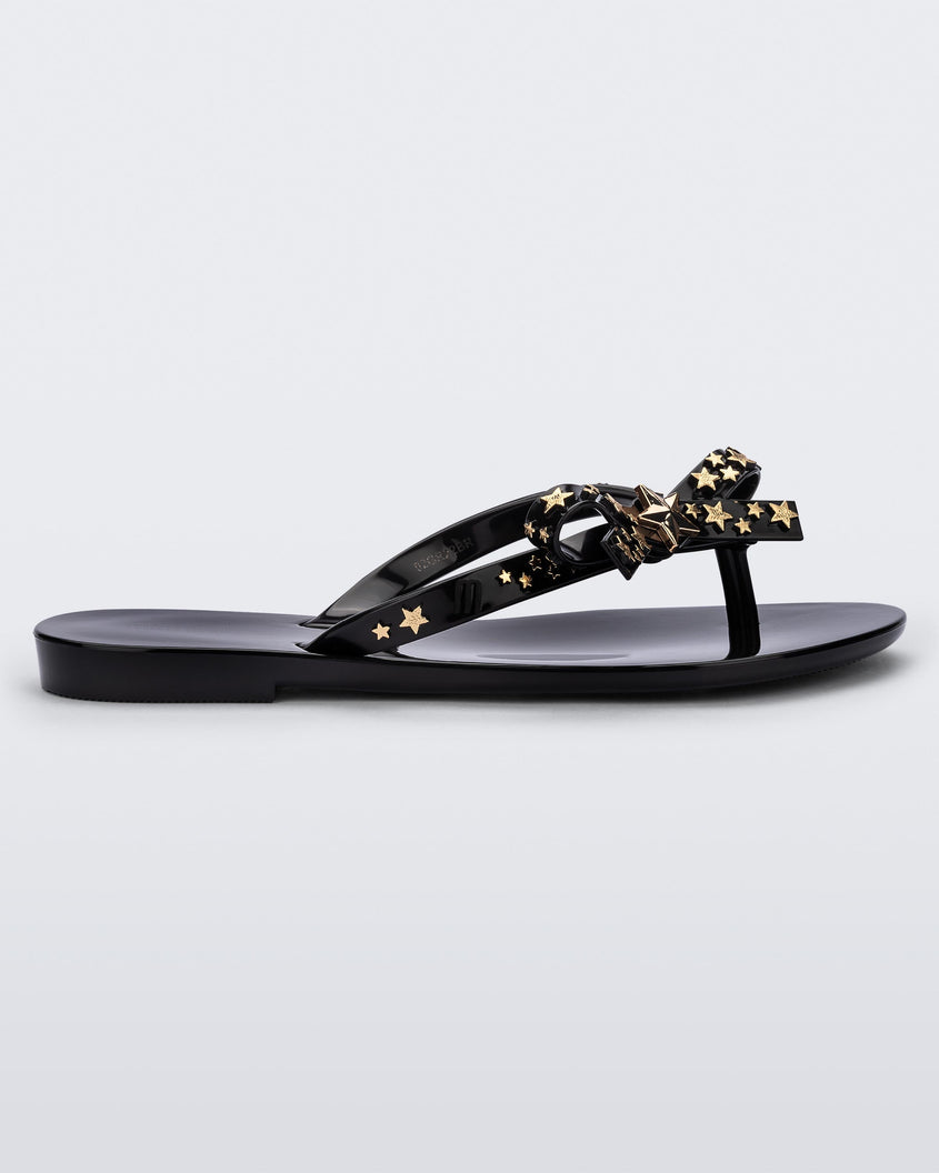 Side view of a black Mini Melissa Harmonic Stars flip flops with a bow and gold star details on the straps.