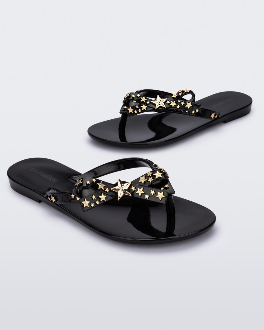 An angled top and side view of a pair of black Mini Melissa Harmonic Stars flip flops with a bow and gold star details on the straps.