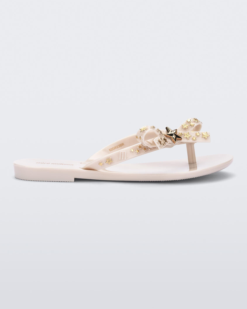Side view of a beige Mini Melissa Harmonic Stars flip flops with a bow and gold star details on the straps.