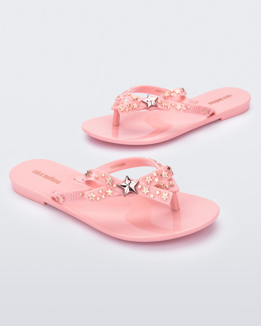 An angled side and front view of a pair of pink Mini Melissa Harmonic Stars flip flops with a bow and gold star details on the straps.