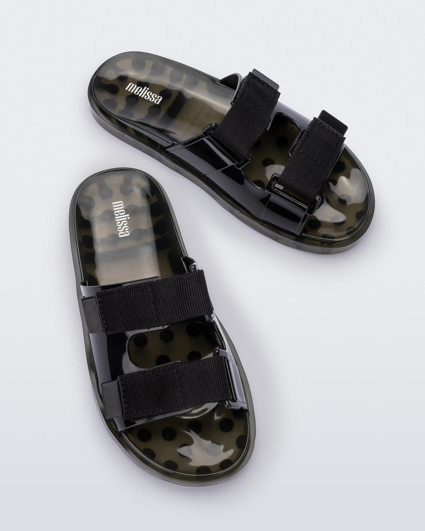 An angled top view of a pair of green/black Melissa Brave Wide Slides with a black base, black velcro straps and a translucent green insole.