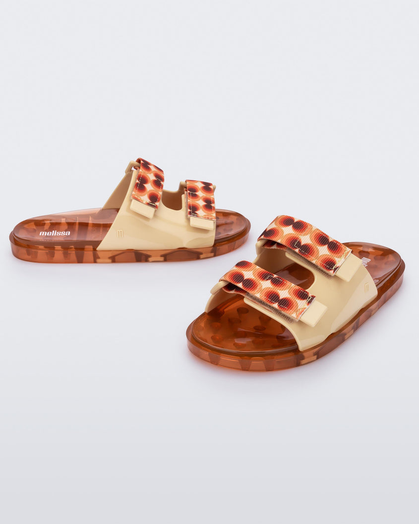 An angled front and side view of a pair of yellow/orange Melissa Brave Wide Slides with a yellow base, red and orange circle print velcro straps and a translucent orange insole.