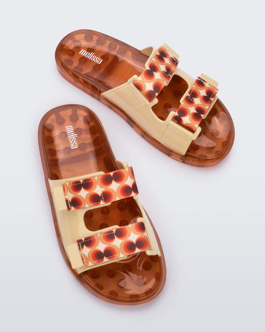 Top view of a pair of yellow/orange Melissa Brave Wide Slides with a yellow base, red and orange circle print velcro straps and a translucent orange insole.