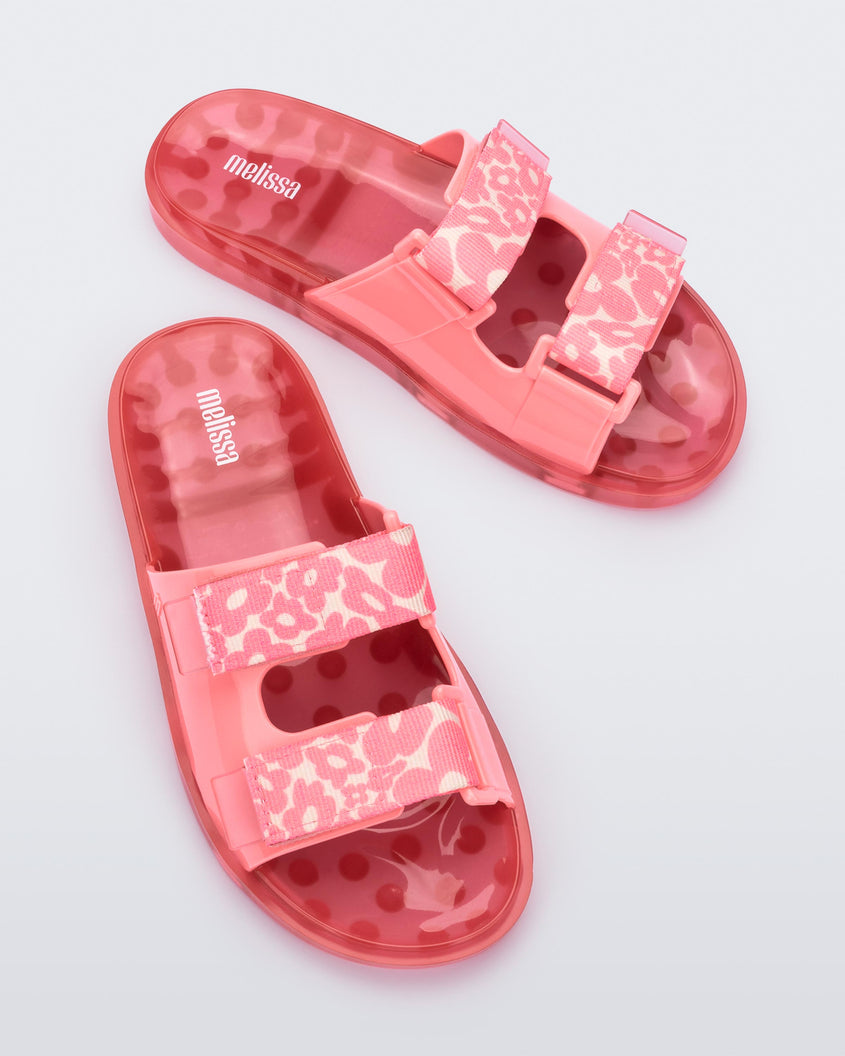 Top view of a pair of pink Melissa Brave Wide slides with a pink base, pink and yellow floral print velcro straps and a translucent pink insole.