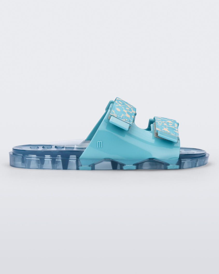 Side view of a blue multicolor Melissa Brave Wide Slide with a blue base, blue and green floral print velcro straps and a translucent blue insole.