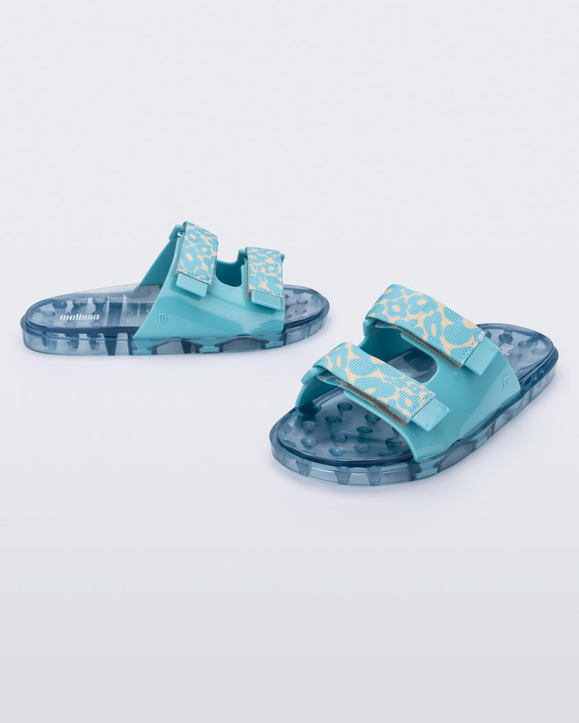 An angled front and side view of a pair of blue multicolor Melissa Brave Wide Slides with a blue base, blue and green floral print velcro straps and a translucent blue insole.