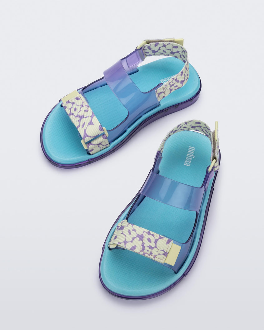 Top view of a pair of lilac Melissa Brave Papete sandals with a blue insole, purple and green flower print and transparent lilac straps and a velcro back.