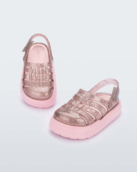 An angled front and top view of a pair of pink/glitter Mini Melissa Sunday sandals with a glitter base, closed toe front and pink sole.