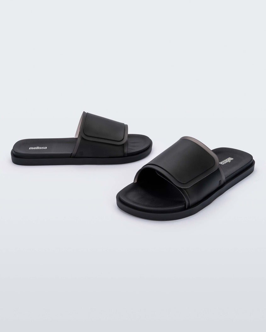 An angled front and side view of a pair of black Melissa Brave Slides with a black velcro top strap and translucent black insole.
