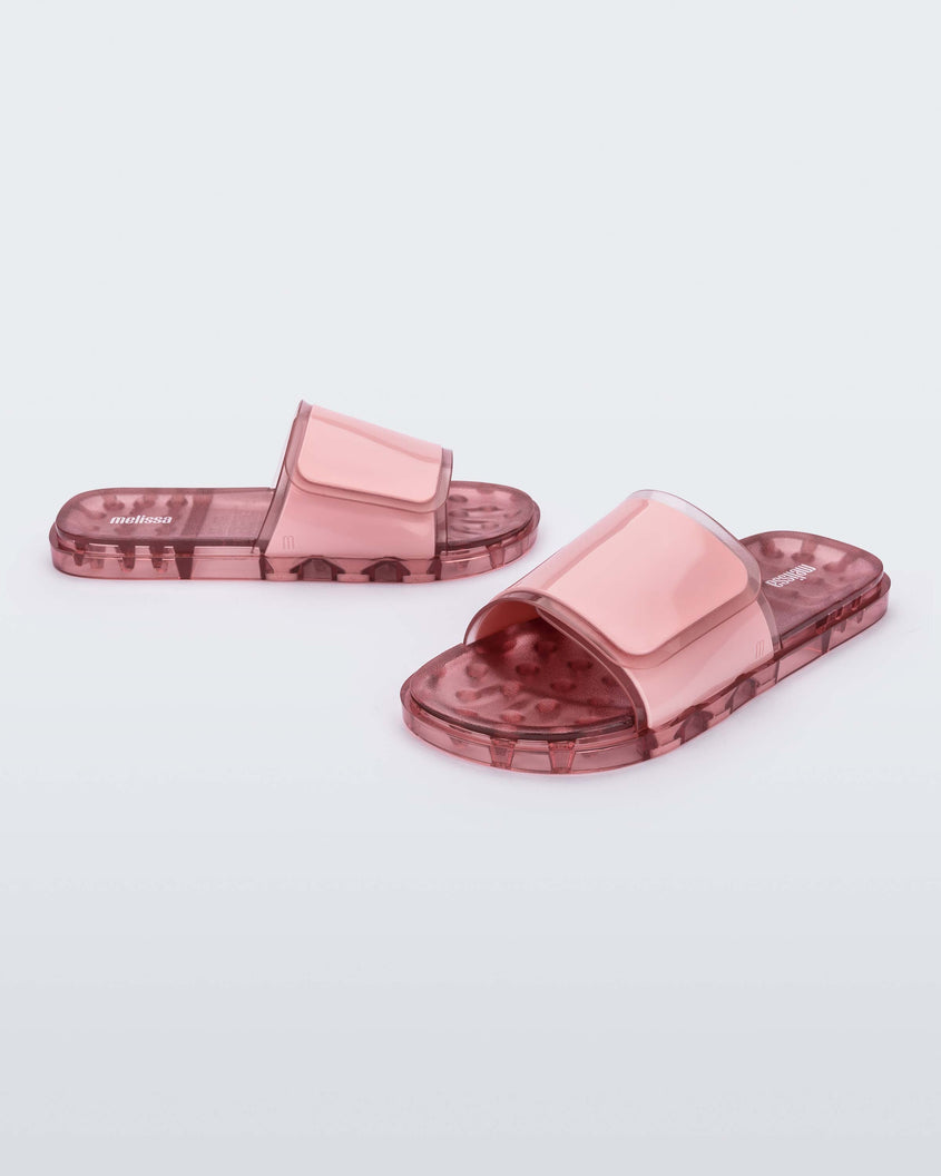 An angled front and side view of a pair of pink Melissa Brave Slides with a pink velcro top strap and translucent pink insole.