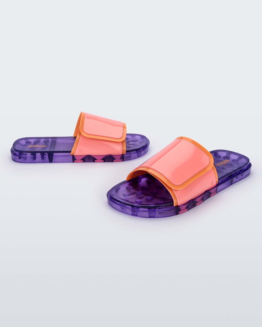 An angled front and side view of a pair of lilac/pink Melissa Brave Slides with a pink and orange velcro top strap and translucent lilac insole.