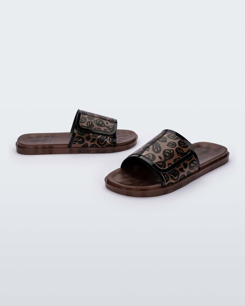 An angled front and side view of a pair of brown / black Melissa Brave Slide slides with a black and brown patterned velcro top strap and translucent brown insole.