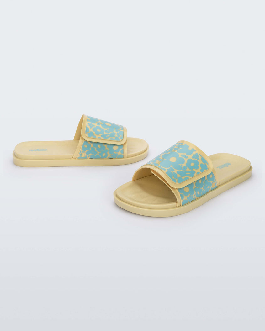 An angled front and side view of a pair of yellow/blue Melissa Brave Slides with a yellow and blue floral patterned velcro top strap and yellow insole.