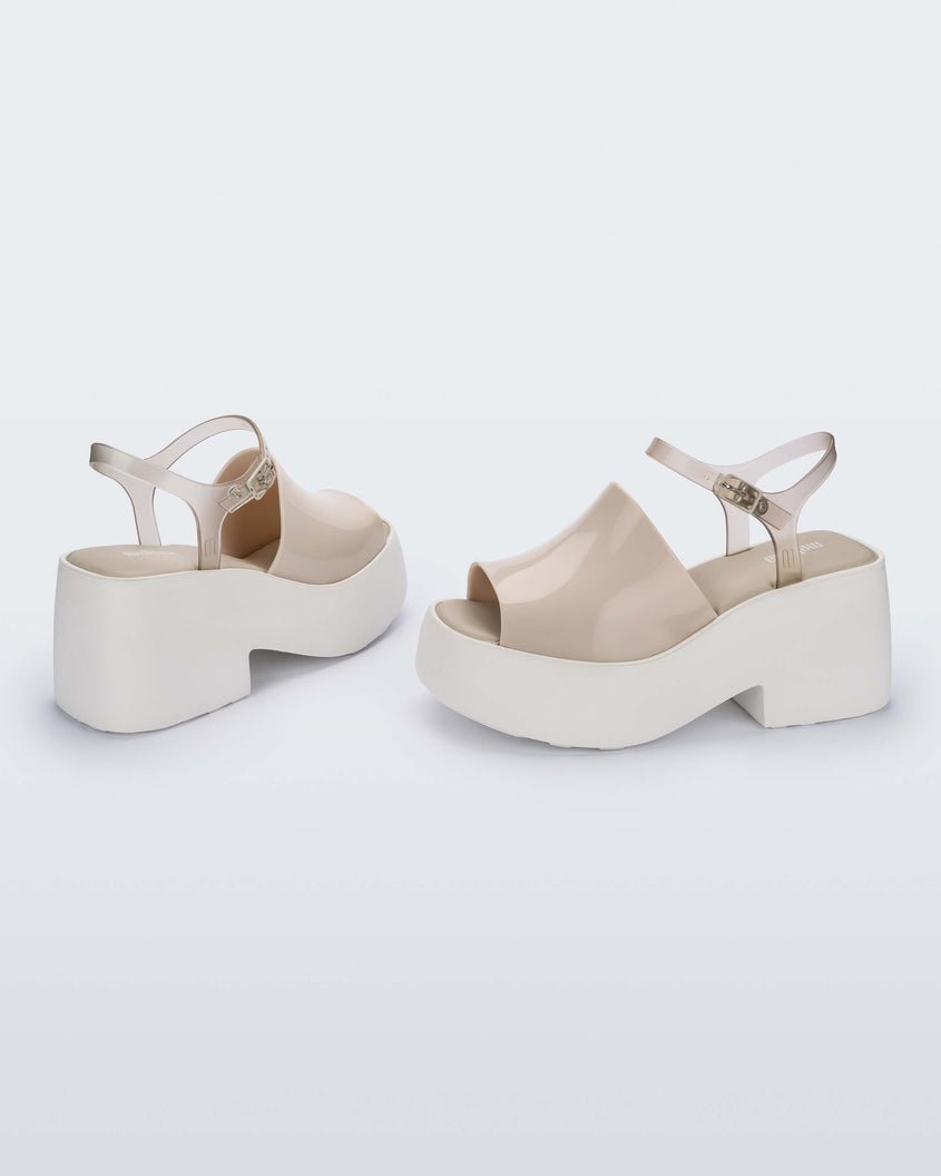 A side and back view of a pair of White/Beige Melissa Pose platform sandals with a beige front strap, a clear beige ankle strap and a white sole.