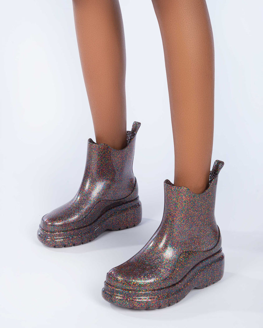 A model's legs wearing a pair of clear glitter Melissa Grip multicolor glitter short rain boots.