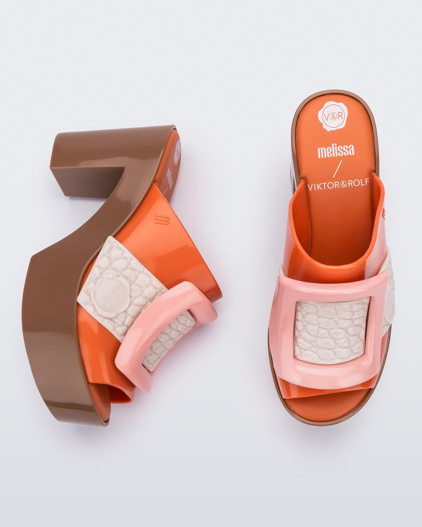 Top and side view of a pair of orange/brown Melissa Buckle Up heeled mules with ivory strap and pink buckle.