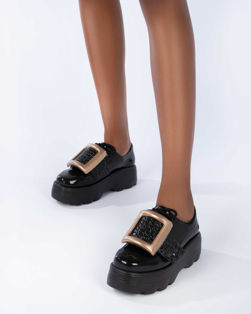 A model's legs wearing a pair of black Melissa Buckle Up Kick Off platform sneakers with a gold buckle.