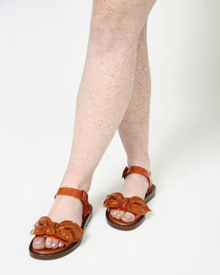 A model's legs wearing a pair of Brown/Orange Melissa Tie Mar Sandals with an orange/brown base, an ankle strap and a front strap with a beige bow on the front.