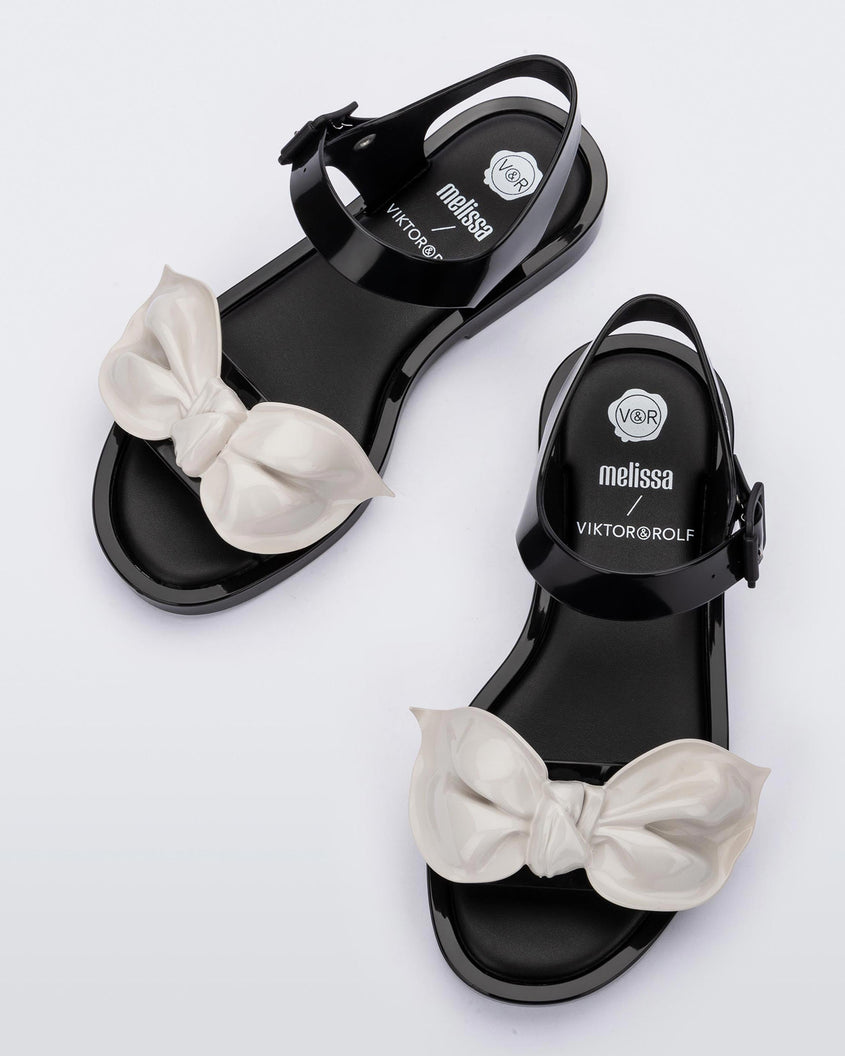 Top view of a pair of Melissa Tie Mar sandals in black with buckle ankle strap and beige 3D bow detail on front straps