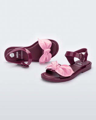 An angled front and top view of a pair of red/pink Melissa Tie Mar Sandals with a red base, an ankle strap and a front strap with a pink bow on the front.