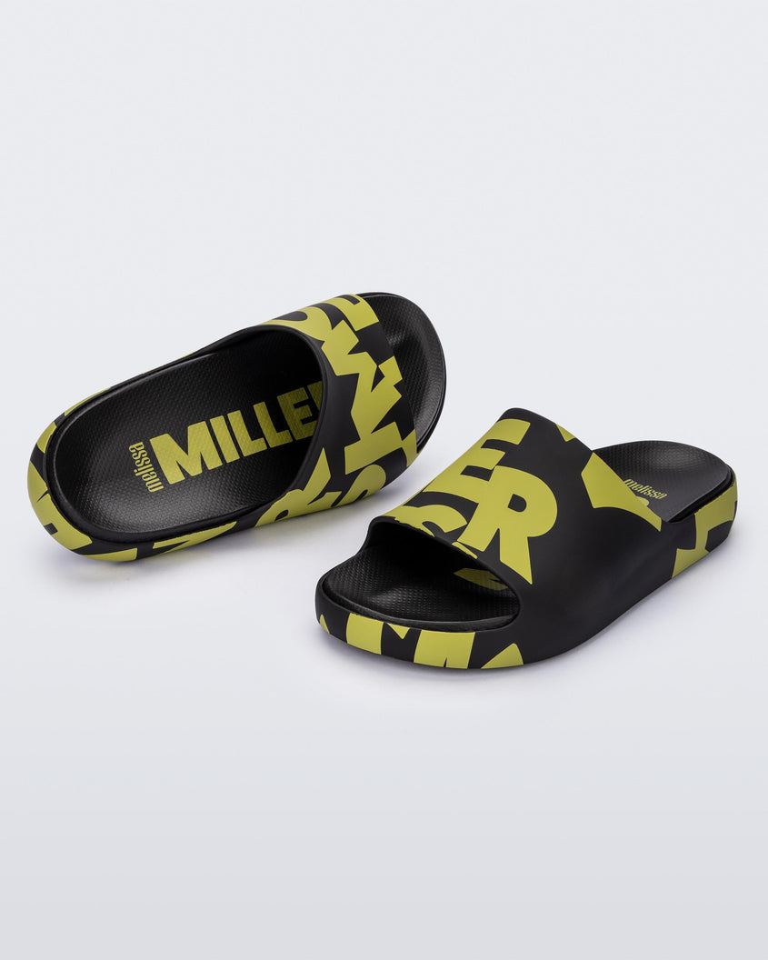 An angled top and front view of a pair of Black & Kiwi Logo Scramble Melissa Cloud slides with a green simon miller letter scrambled text on the base and a black insole reading in green 