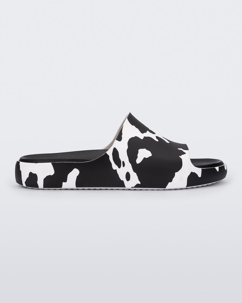 Side view of a pair of black and white cow print Melissa Cloud slides with a black insole reading in blue "Melissa Miller" and "Melissa Simon" on different shoes with a cow print base.