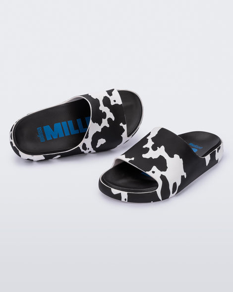 An angled front and top view of a pair of black and white cow print Melissa Cloud slides with a black insole reading in blue 