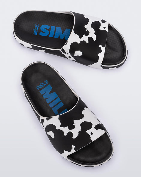 An angled top and side view of a pair of black and white cow print Melissa Cloud slides with a black insole reading in blue 
