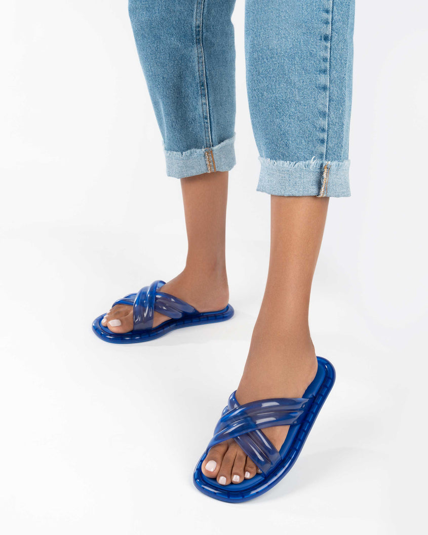A model's legs wearing a pair of transparent blue Melissa Cali slides with cross straps.