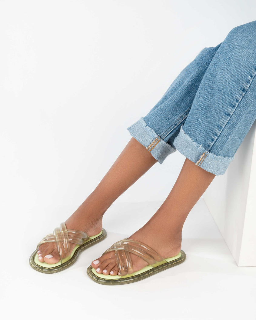 A model's legs wearing a pair of transparent green Melissa Cali slides with cross straps.
