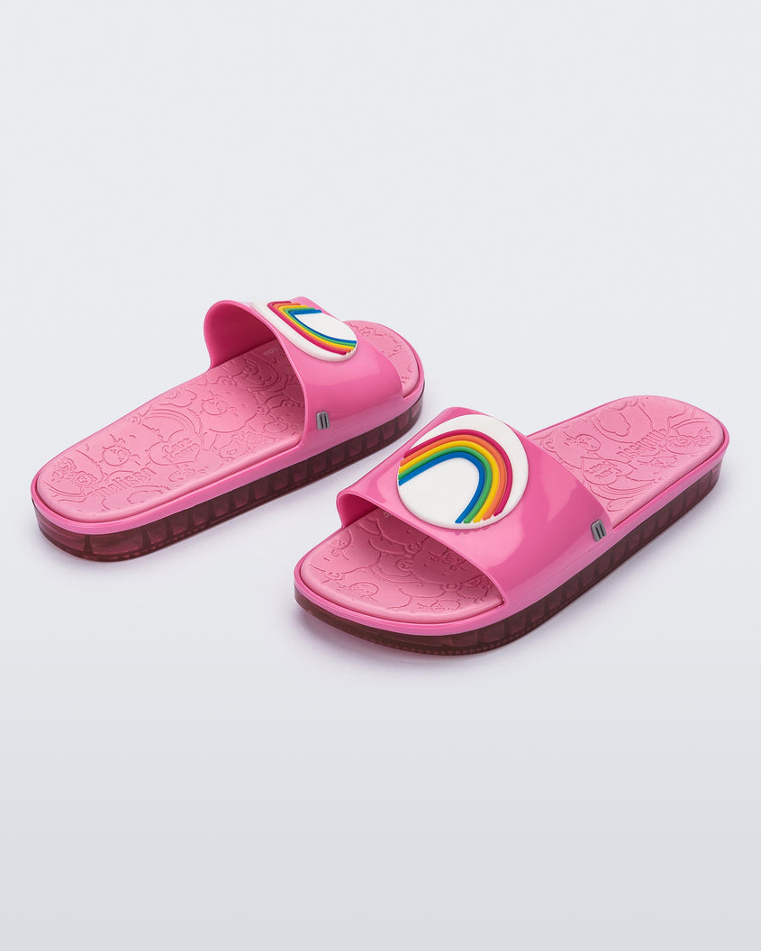 An angled front and side view of a pair of pink Melissa Beach slides with a rainbow on a white circle on the top strap, and a care bear patterend pink insole.