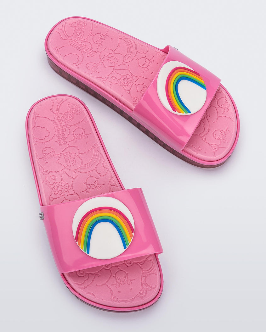 Top view of a pair of pink Melissa Beach slides with a rainbow on a white circle on the top strap, and a care bear patterend pink insole.