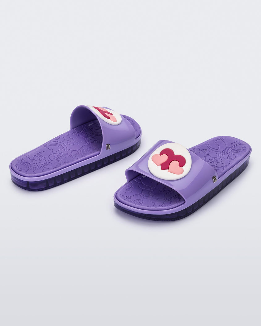 An angled front and side view of a pair of lilac Melissa Beach slides with 3 pink hearts on a white circle on the top strap, and a care bear patterend lilac insole.