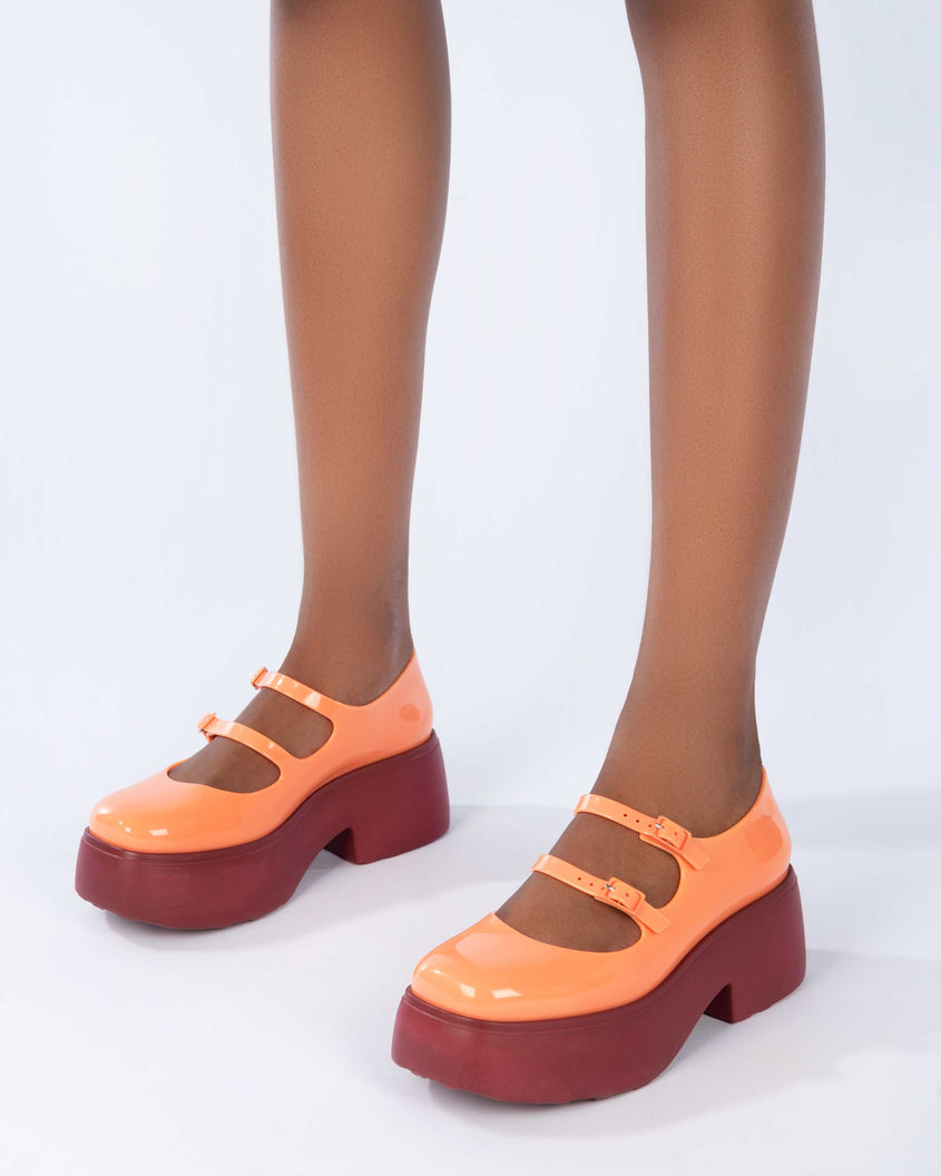A model's legs wearing a pair of orange Melissa Farah platform shoes, with a brown sole and two orange straps fastened by orange buckles.